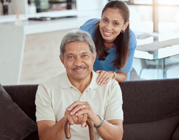 When Coverage Changes: How To Provide Stable Home Rehab Nursing Assistant in blue scrubs standing and smiling behind a gray-haired Hispanic man sitting on a couch with a cane.