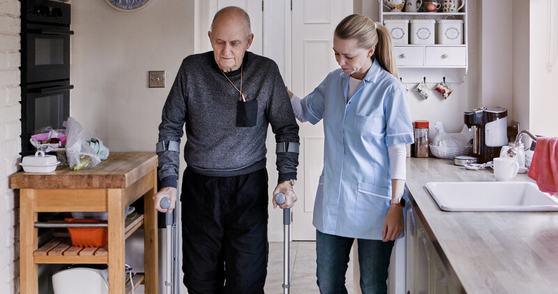 Nurse help elderly man in crutches in home kitchen, medical rehabilitation and recovery. Caregiver, person with a disability walk and support, health or physiotherapy, healing injury or accident pain. Brain Injury Recovery: How to Support Rehab at Home
